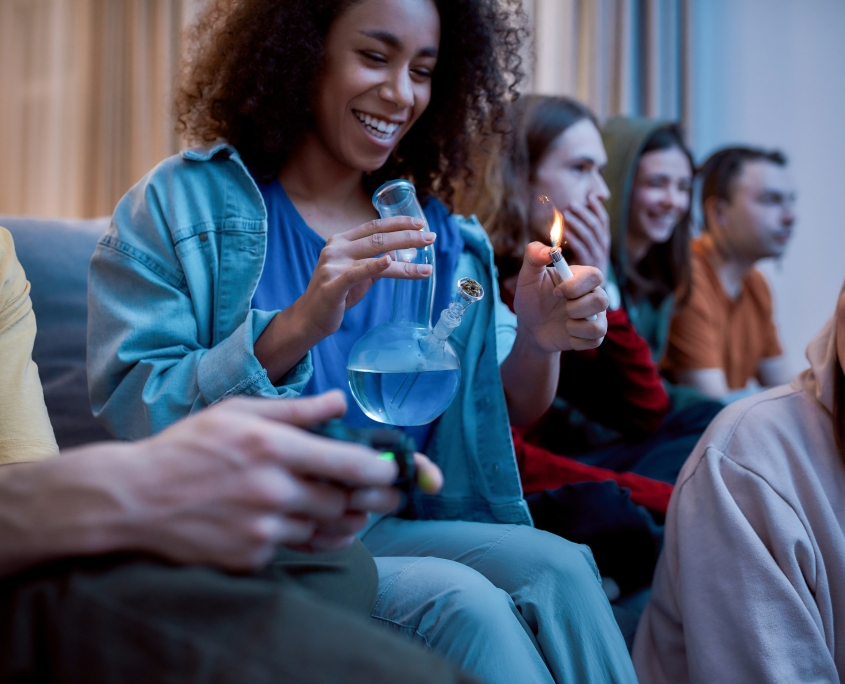 Friends smoking weed at home. Young afro american girl lighting marijuana in the glass bong, relaxing with friends on the sofa at home. Young people playing video games. Weed, cannabis legalization